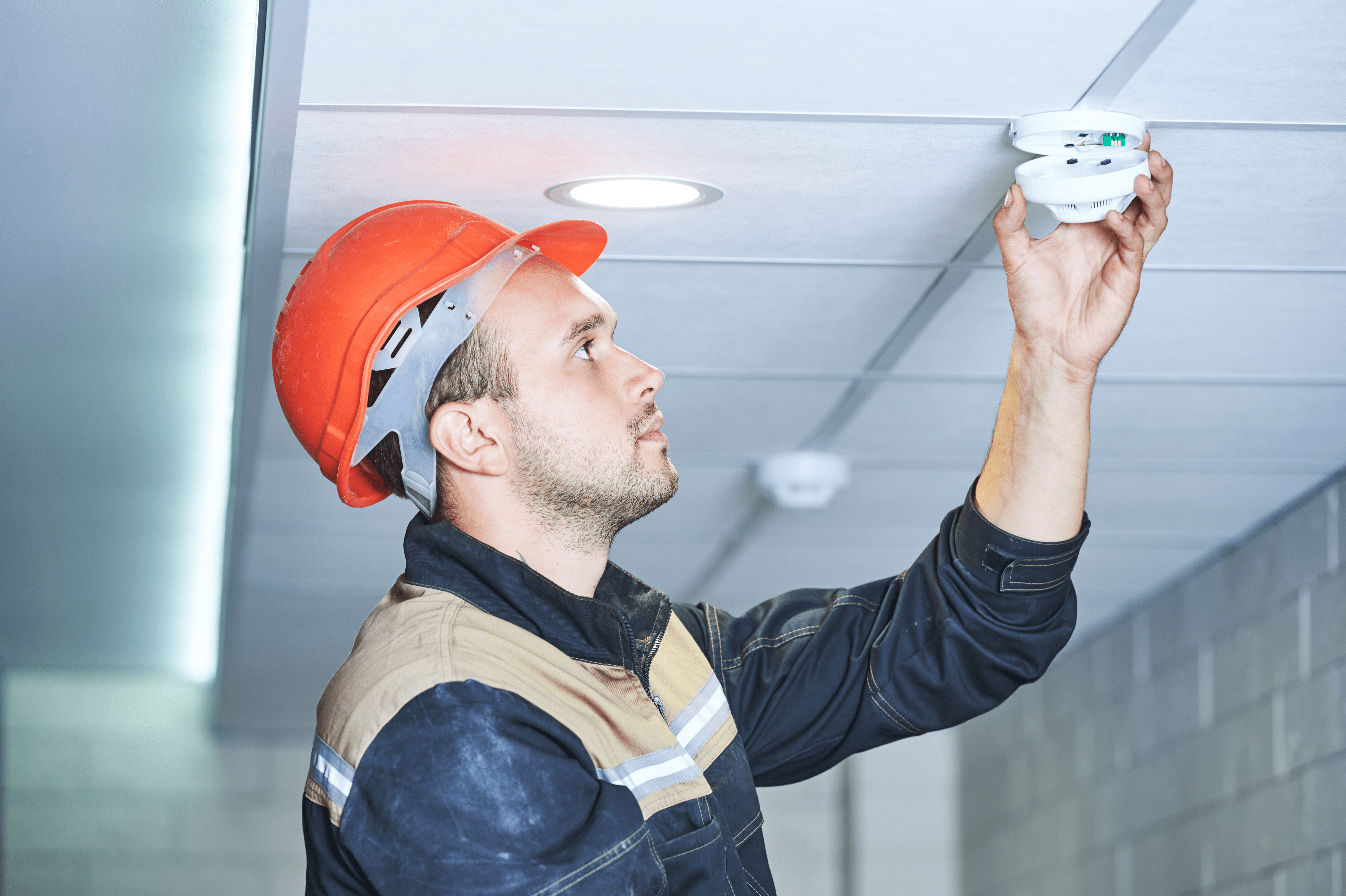 A man reaching up toward a fire alarm which is hanging loose, he has a hard hat and vest on and is looking at it while installing it.